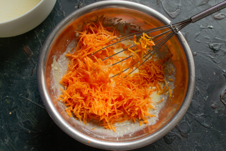 Incorporating Freshly Grated Carrots into the Batter A hand adding a pile of finely grated orange carrots to the tvorog mixture in a bowl.