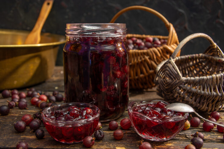 A glass jar filled with vibrant red gooseberry jam, with a spoon resting beside it.