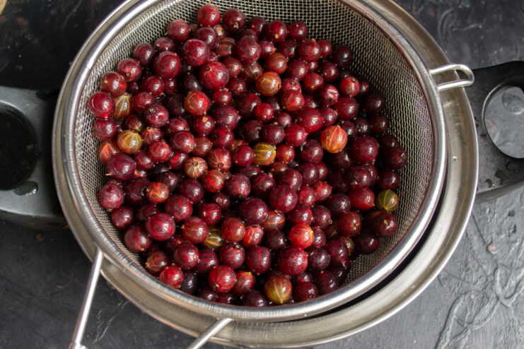 Red gooseberries draining in a metal sieve over a bowl.
