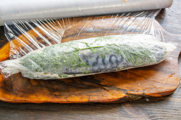 Step 5: How to Tightly Wrap the Mackerel Roll in Plastic Film A mackerel roll being tightly wrapped in clear plastic wrap.