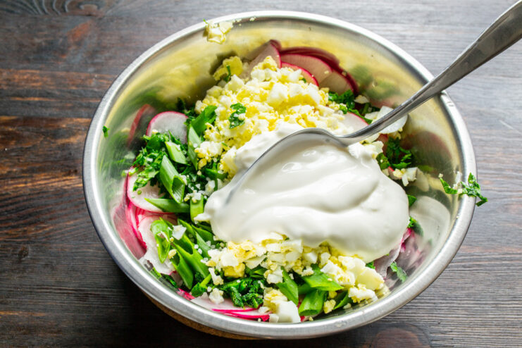 Creating the Creamy Dressing with Smetana or Yogurt A spoon adding a dollop of smetana (sour cream) to the salad ingredients in a bowl.