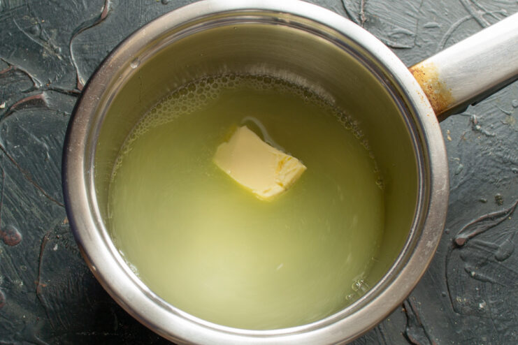 Step 1: Melting butter with milk whey, sugar, and salt for the dough. A saucepan on a stove with milk whey, sugar, salt, and a piece of melting butter.