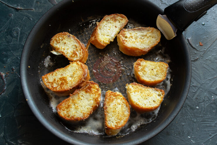 Frying baguette slices in butter until golden brown Baguette slices being fried to a golden brown color in a pan with melted butter.