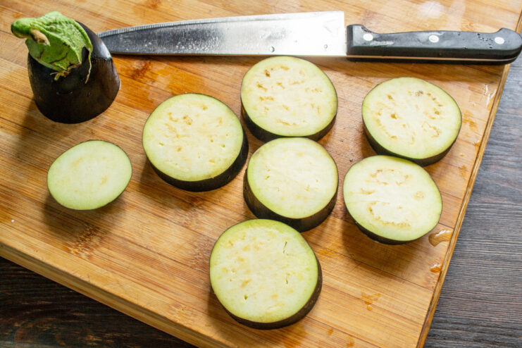 Slicing eggplant into 1.5 cm thick rounds for even baking. A hand slicing a fresh eggplant into thick rounds on a cutting board.