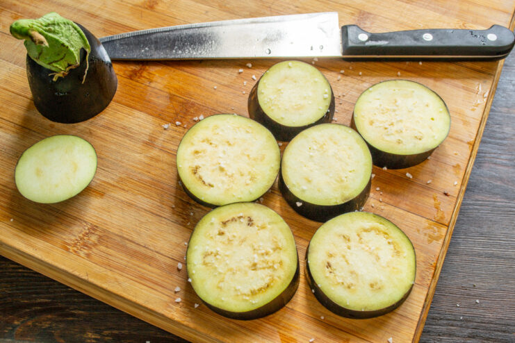 Salting the eggplant slices before baking to enhance flavor. Sliced eggplant rounds on a cutting board being sprinkled with salt.