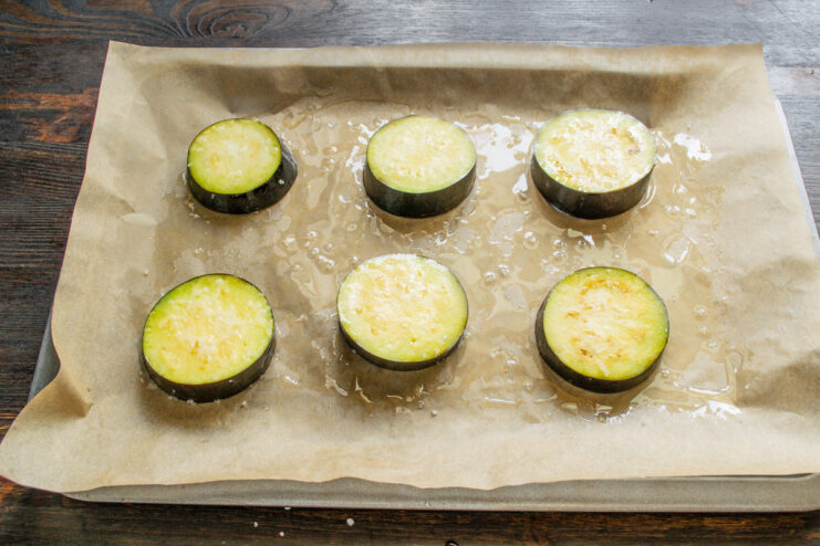 Coating eggplant slices with olive oil on both sides for a golden finish. Eggplant slices coated in olive oil arranged on a parchment-lined baking sheet.