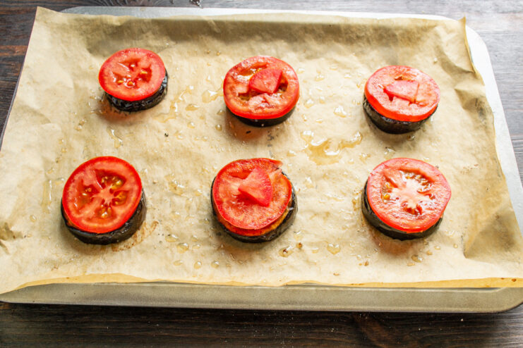 Layering fresh tomato slices on top of the baked eggplant. Baked eggplant slices on a baking sheet, each topped with a fresh tomato slice.