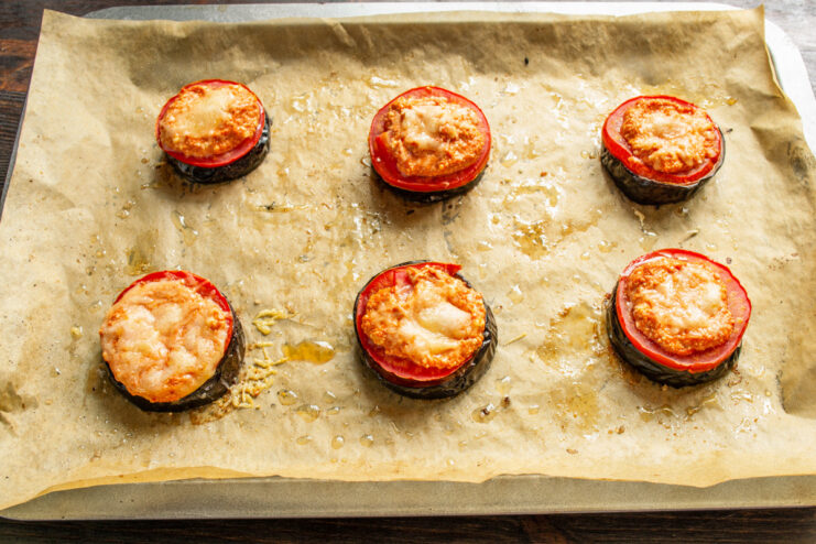 Baking the eggplant stacks again until the cheese is melted and bubbly. A baking sheet with eggplant, tomato, and cheese stacks going into the oven.