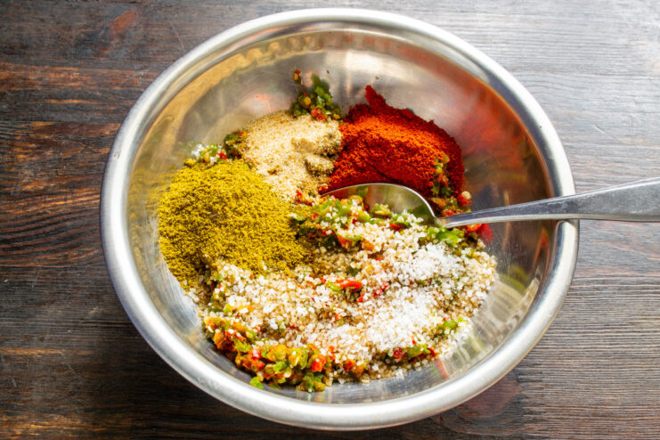 Adding Spices to the Adjika Mixture A bowl of minced vegetable paste with various spices being added on top.