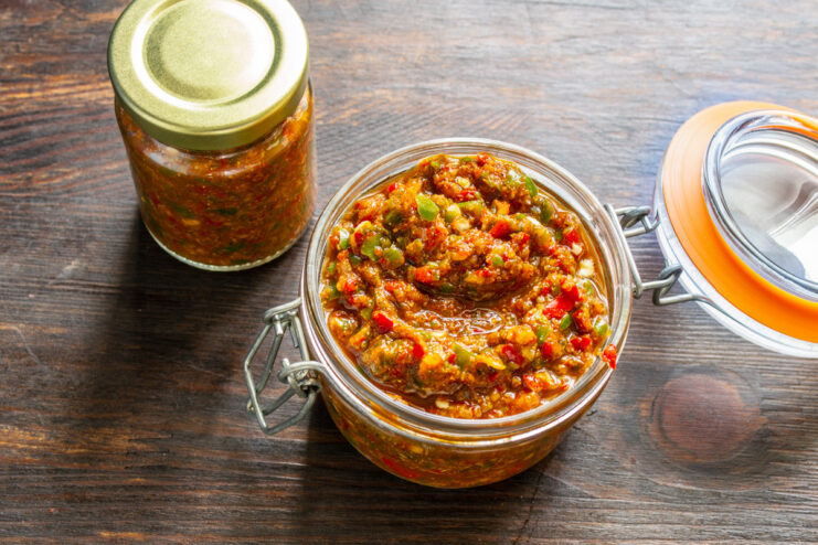 Storing Homemade Adjika in Jars A hand using a spoon to pack bright red adjika into a clean glass jar for storage.