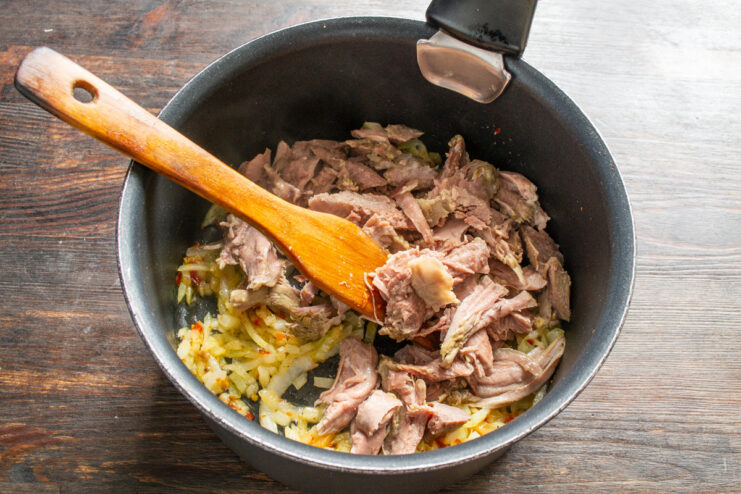 Adding shredded cooked meat to the sautéed onions. Shredded cooked meat being added to the pot with sautéed onions.