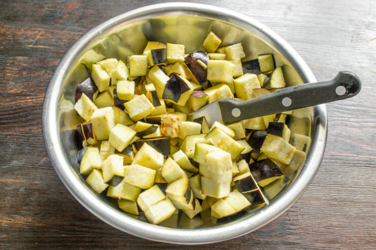 Preparing the Eggplant: Dicing into Half-Inch Cubes A hand holding a knife dicing a purple eggplant into small cubes on a wooden cutting board.