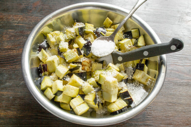 Salting the Eggplant to Draw Out Bitterness and Moisture Diced eggplant in a glass bowl being sprinkled with coarse sea salt from above.