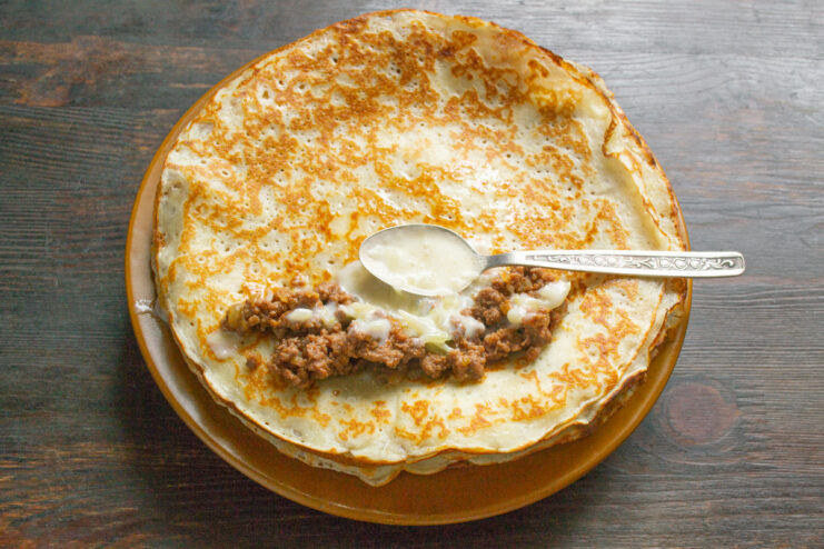 Assembling the Blinchiki with Meat Filling and Sauce A cooked blinchik laid flat with a spoonful of meat filling and cream sauce on top.