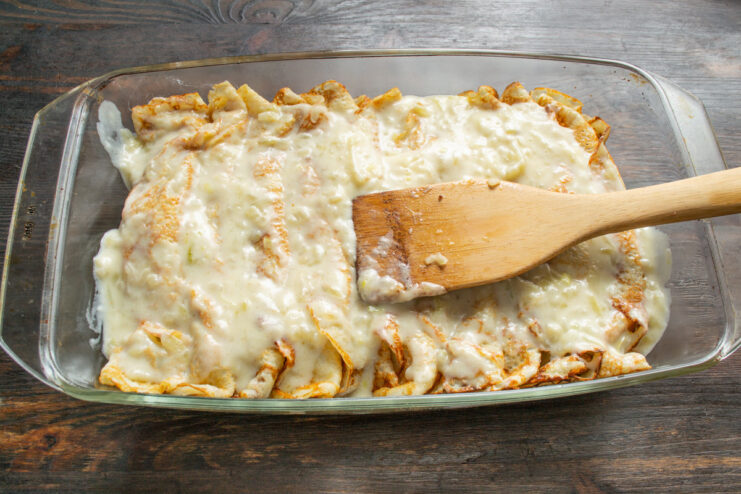 Pouring Cream Sauce Over Blinchiki Before Baking Pouring a rich, creamy sauce over the arranged blinchiki in a baking dish.