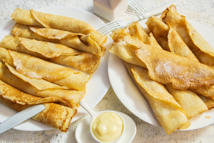 A stack of rolled blinchiki, or thin pancakes, filled with lemon custard and garnished with powdered sugar on a white plate.