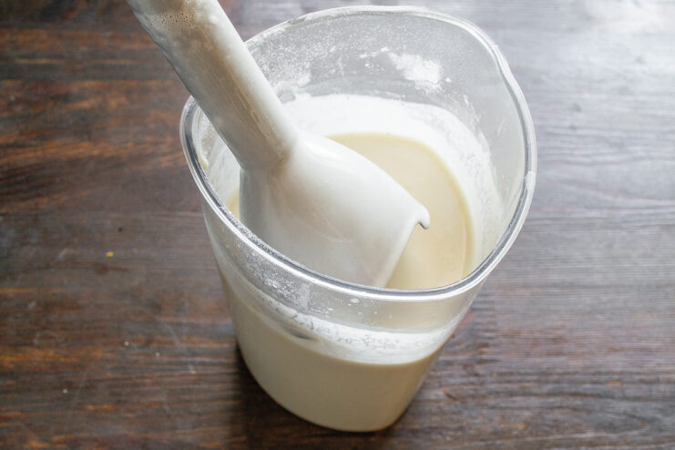 An immersion blender mixing pancake batter in a tall container with ingredients like egg, flour, and milk.