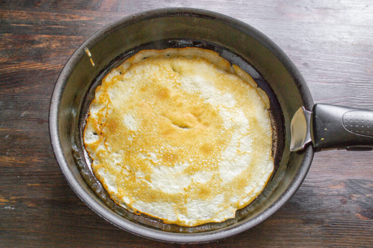 A thin, golden-brown pancake being cooked in a black frying pan.