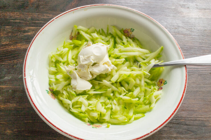 Adding Cream Cheese to the Zucchini Batter A spoonful of soft cream cheese being added to a bowl of grated zucchini.