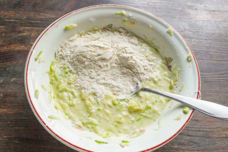 Adding Flour to Thicken the Zucchini Batter Adding a spoonful of wheat flour to the zucchini pancake batter in a bowl.