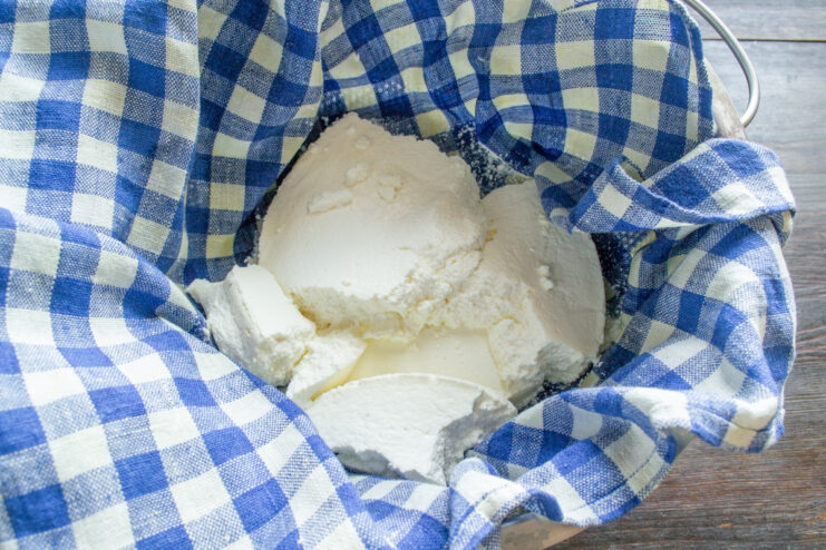 Draining Homemade Tvorog in the Refrigerator Homemade tvorog wrapped in a white cloth, placed in a colander over a bowl to drain excess whey.