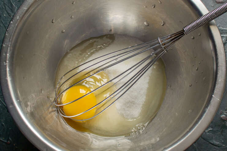 Whisking egg and sugar together until pale and fluffy for the bun batter. A glass bowl with an egg and sugar being whisked together with a metal whisk.