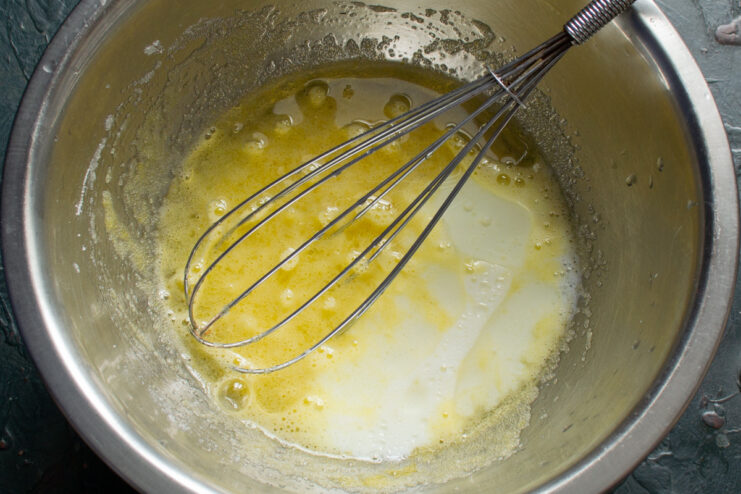 Combining wet ingredients: adding milk and melted butter to the egg mixture. Pouring a milk and butter mixture from a small saucepan into a glass bowl with whisked eggs and sugar.