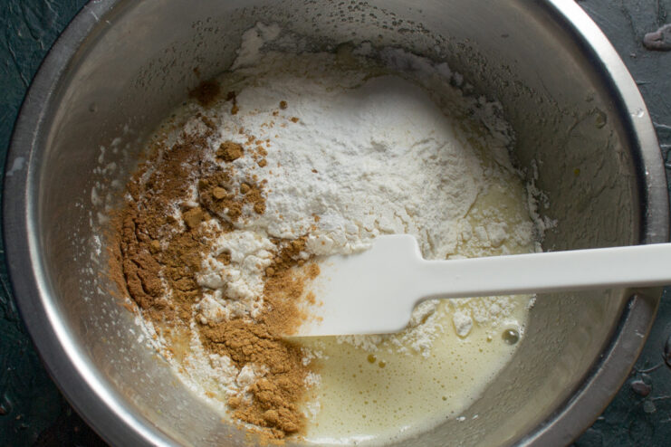 Adding all-purpose flour to the wet ingredients to form the batter. Sifting all-purpose flour into a glass bowl containing the wet ingredients for the apple buns.