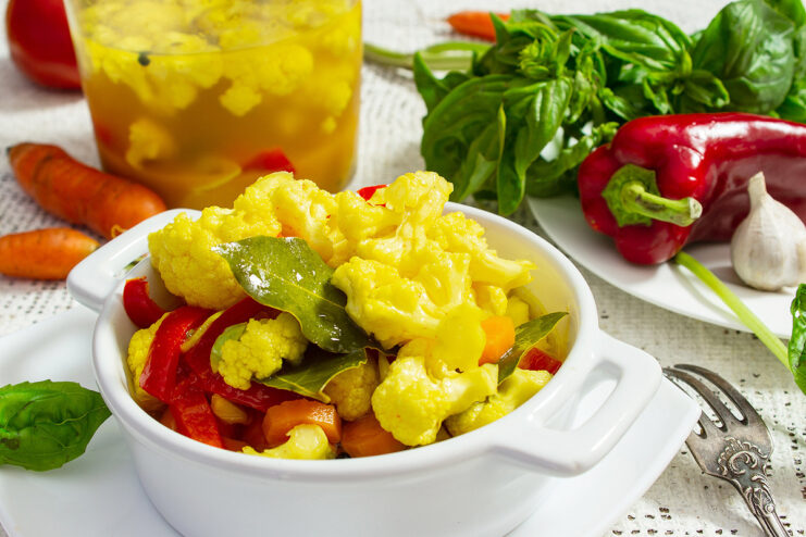 A glass bowl filled with yellow marinated cauliflower florets, carrot slices, and red pepper strips.