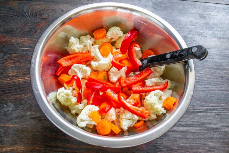 Step 3: Combining the vegetables before marinating A white bowl containing cauliflower florets, carrot slices, and freshly added strips of red bell pepper.