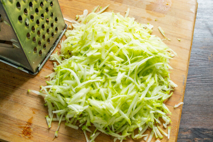 Grating Zucchini for the Draniki Batter Grating a peeled zucchini on a coarse box grater into a bowl.