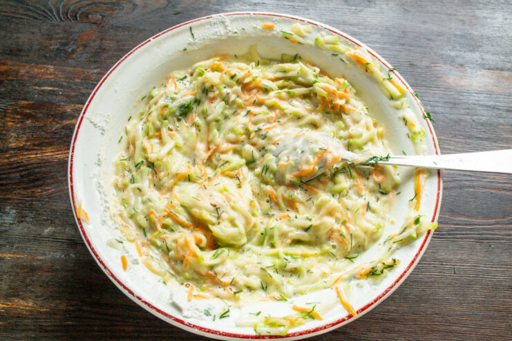 Mixing the Draniki Batter Until Well Combined A spoon mixing the zucchini, carrot, and flour batter in a white bowl.