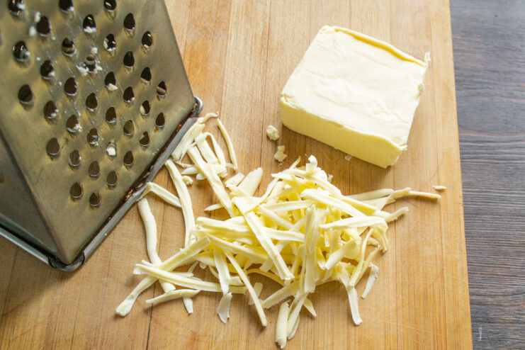 Grating Processed Cheese for the Salad Base A block of processed cheese being grated on a coarse box grater into a clear bowl.