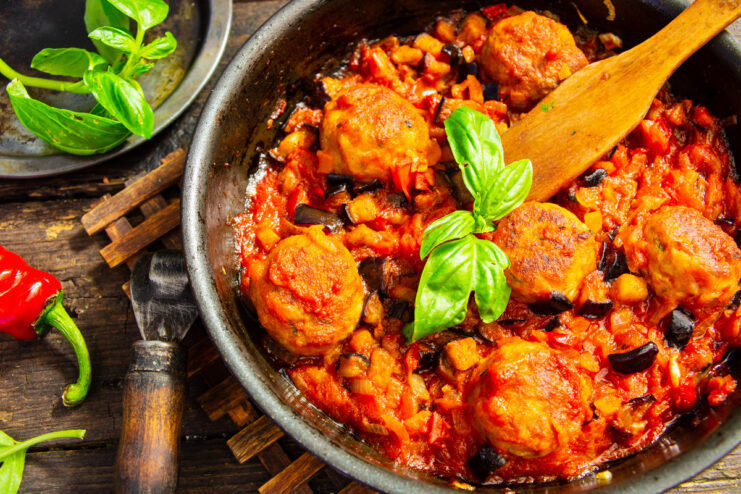 A white bowl filled with meatballs and eggplant in tomato sauce, garnished with fresh herbs.