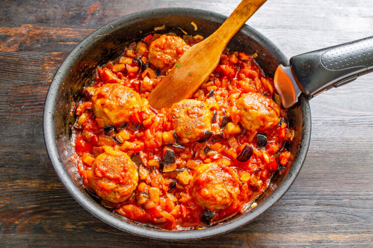 Meatballs and eggplant simmering together in tomato sauce in a covered pan.