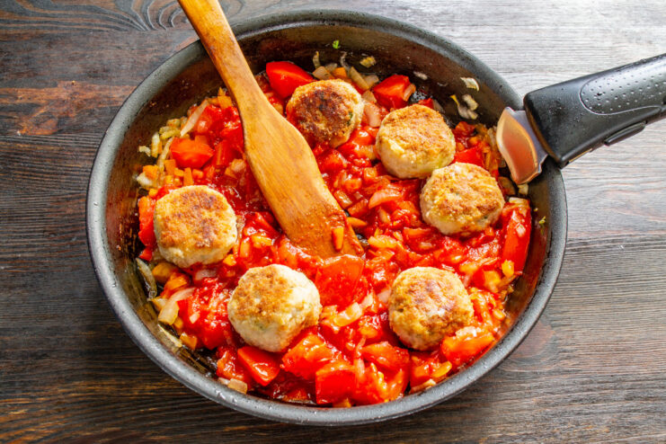 Fried meatballs being added to the simmering tomato sauce in a pan.
