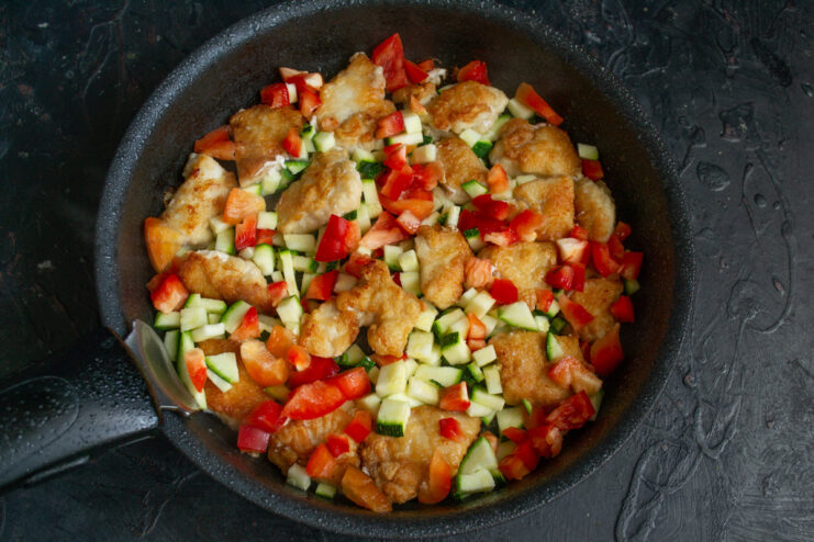 Diced zucchini and bell pepper being added to the skillet with golden-brown chicken pieces.