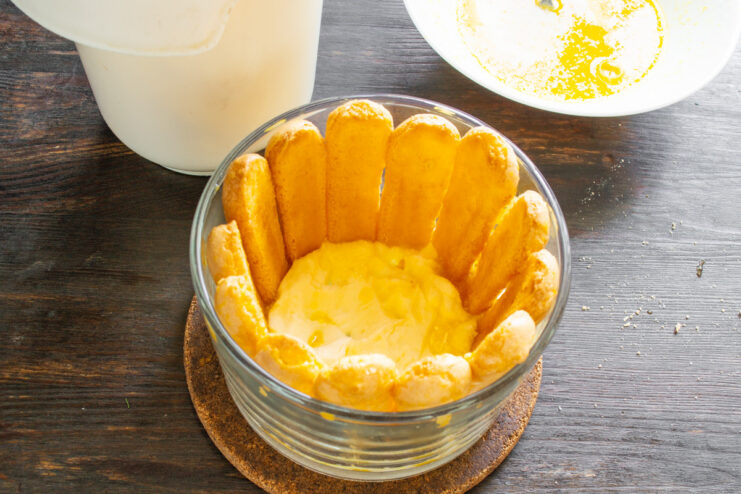 Quickly dip each ladyfinger in the orange juice mixture before layering. A hand dipping a savoiardi biscuit (ladyfinger) into a bowl of orange juice.