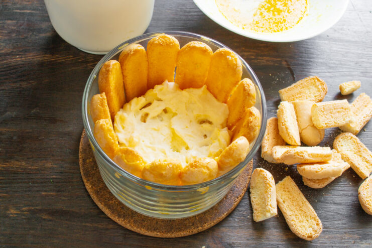 Layering the cake with orange-soaked ladyfingers and mascarpone cream. Assembling the no-bake cake with layers of cream and biscuits in a glass dish.