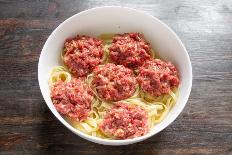 Stuffing the Pasta Nests with Beef Patties A hand placing a small beef patty into the center of a pasta nest in a baking dish.