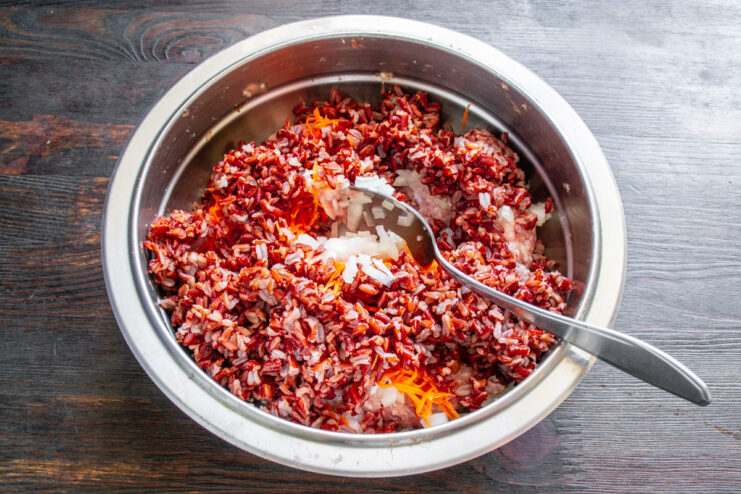 Combining cooked red rice with the meat and vegetable filling. Mixing cooked red rice into a bowl with ground meat, onions, and carrots.