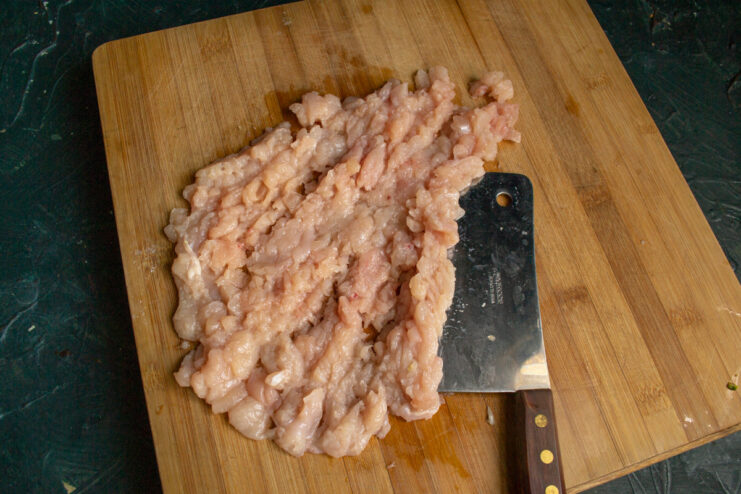 Close-up of chicken breast being finely chopped into mince with a kitchen cleaver on a cutting board.