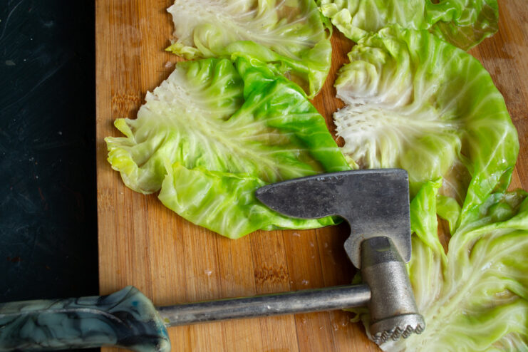 Using a meat mallet to flatten the tough stem of a blanched cabbage leaf on a cutting board.