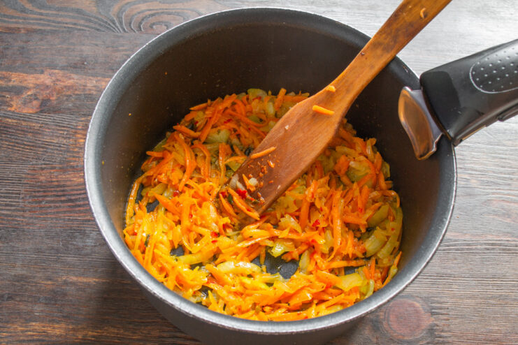 Grated carrots being added to a pot with sautéing onions.