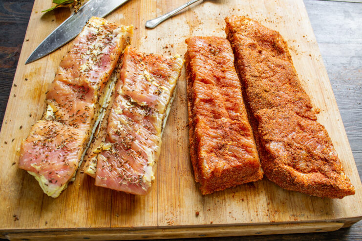 Coating the Pork Belly in Two Different Spice Blends Two pieces of cured pork belly, one coated in a red spice mix and the other in caraway seeds.