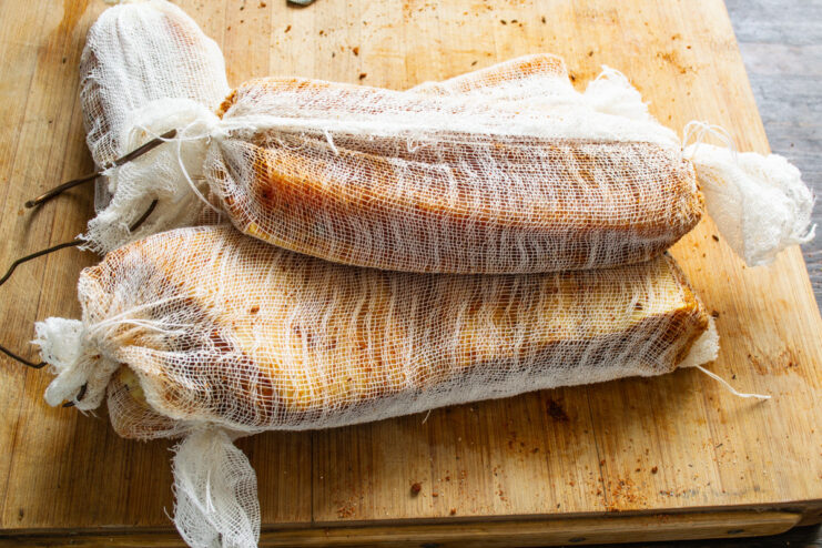 Aging the Spiced Pork Belly in Cheesecloth Two pieces of spice-coated pork belly wrapped and tied in white cheesecloth, ready for aging.