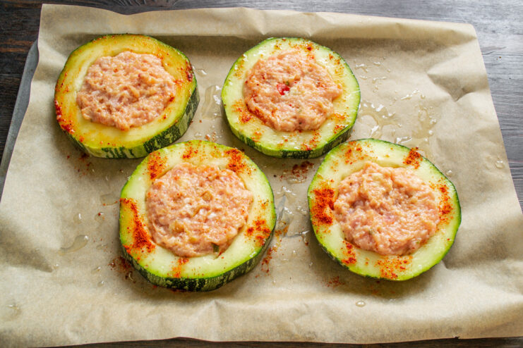 Stuffing the zucchini rings with the prepared minced meat filling. A hand pressing a ball of minced meat filling into a zucchini ring on a baking sheet.