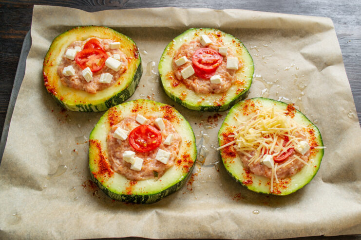 Topping the stuffed zucchini with tomato and feta cheese. A slice of tomato and feta cheese cubes being placed on top of the meat-stuffed zucchini.