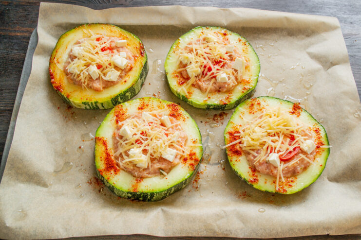 Assembling all the stuffed zucchini rings before baking. A baking sheet full of assembled zucchini rings stuffed with meat and topped with tomato and cheese.