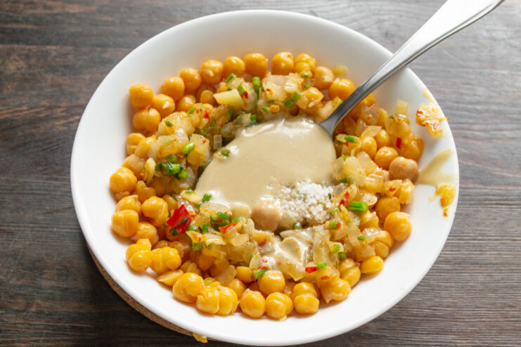 Adding tahini and sea salt to the chickpea mixture for flavor. Adding a spoonful of tahini to the bowl with chickpeas and vegetables.